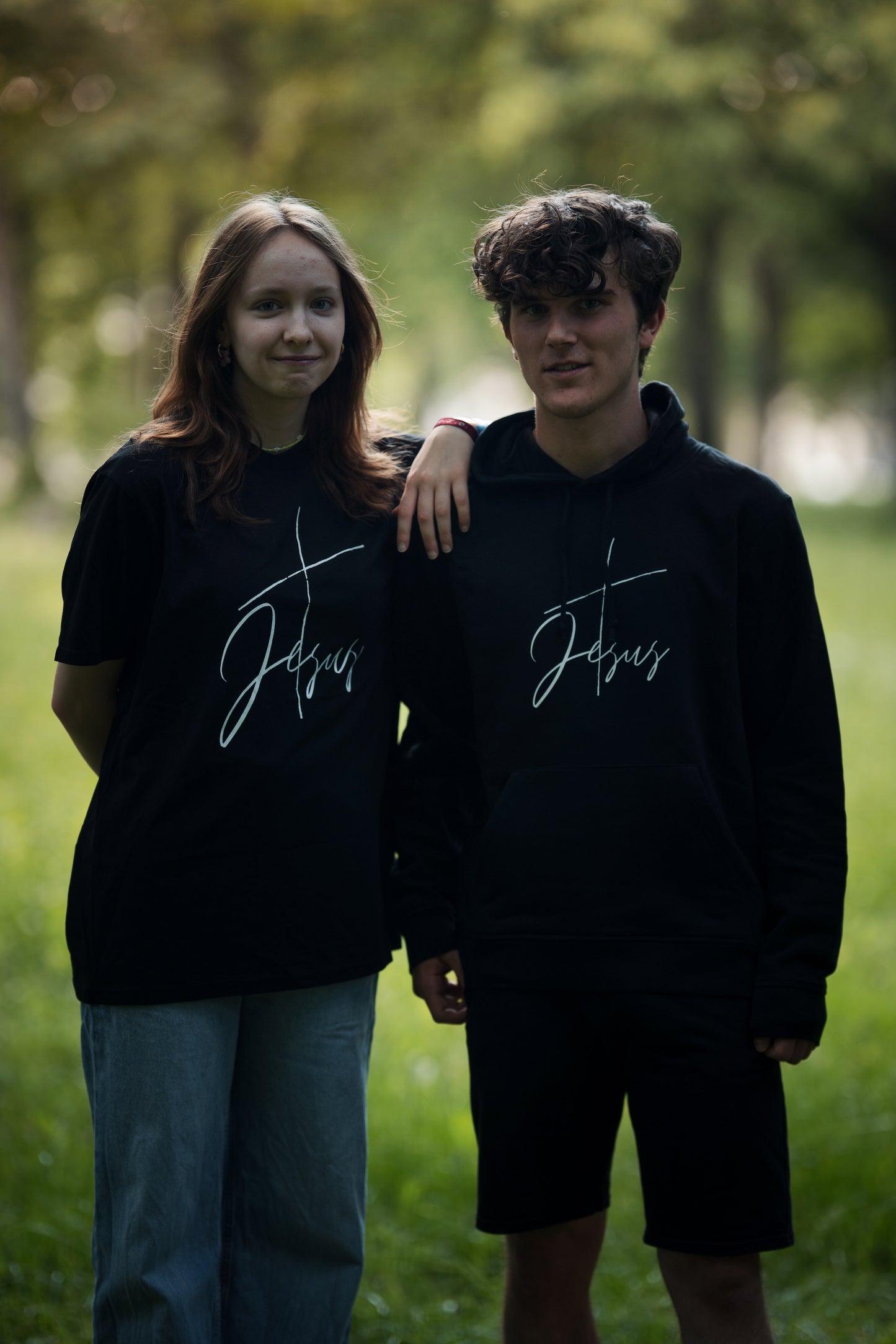 christliches T-Shirt und Hoodie mit einem Unisex-Schnitt an 2 Models in schwarz aus Bio-Baumwolle "Kreuz Jesus" mit einem Kreuz und dem Schriftzug Jesus mitten auf der Brust. Erhältlich in den Größen S bis XXL